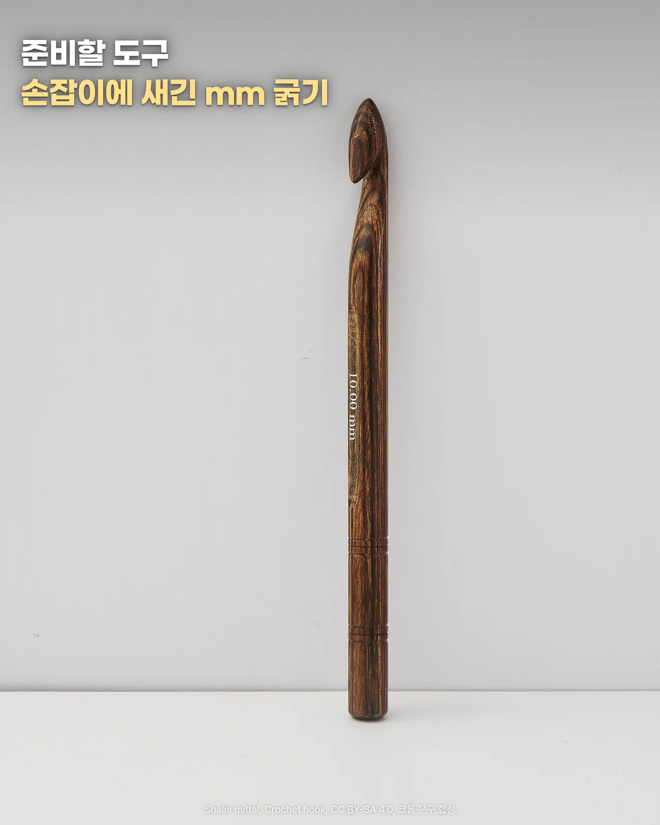 흰 벽 앞 바닥에 세워 둔 갈고리 끝이 위를 향한 나무 코바늘 한 자루, 손잡이에 굵기 10.00 mm가 새겨진 사진에 '준비할 도구', '손잡이에 새긴 mm 굵기' 문구가 있는 이미지