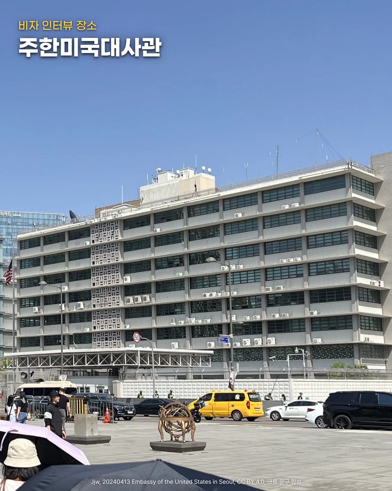 광화문 세종대로에서 바라본 주한미국대사관. 성조기가 게양된 회색 사무동과 정문, 담장이 맑은 하늘 아래 서 있고, 위쪽 하늘에 '비자 인터뷰 장소'와 '주한미국대사관' 글자를 넣은 사진입니다.