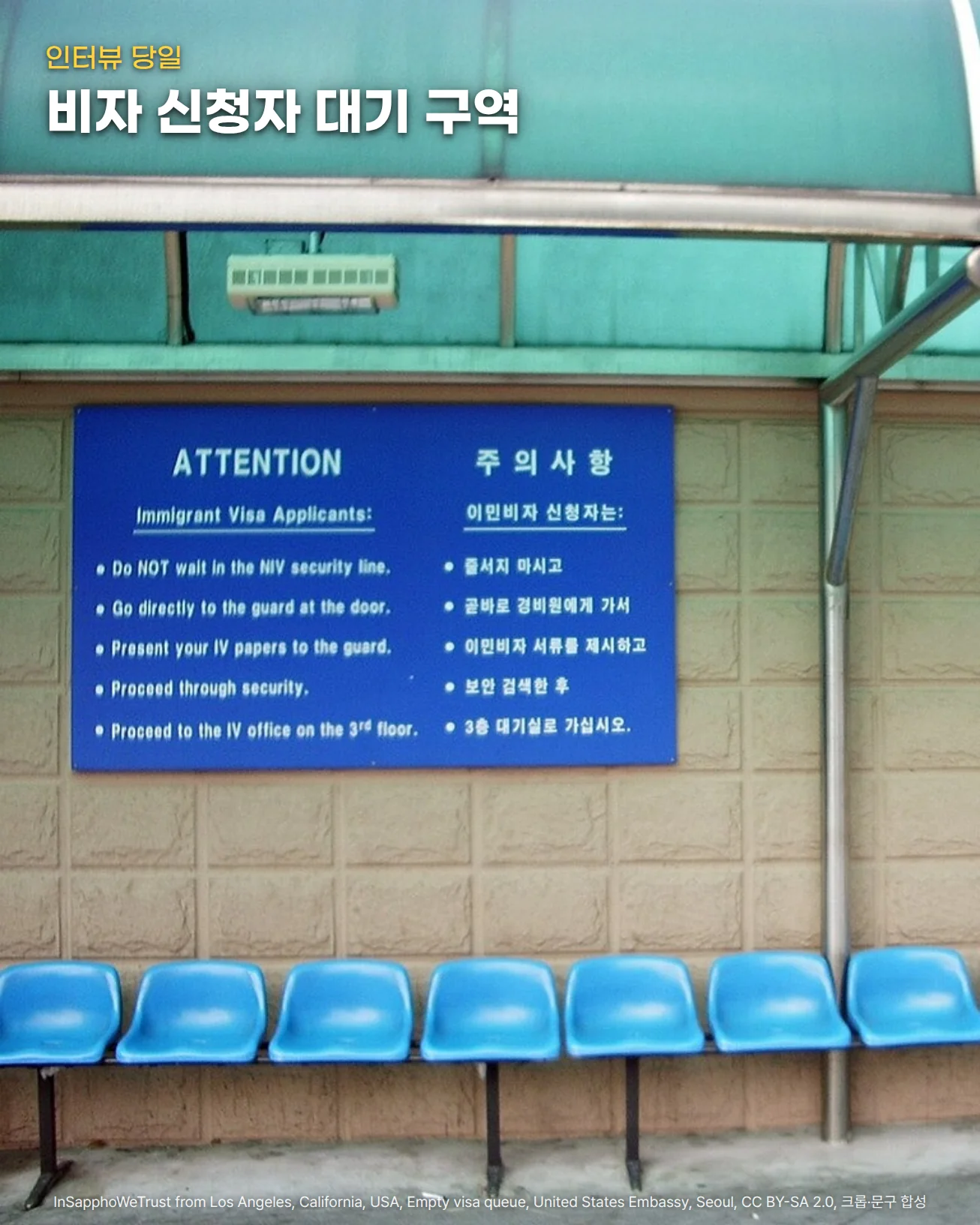 주한미국대사관 앞 비자 신청자 대기 구역. 청록색 차양 아래 담장에 영어와 한국어로 된 파란 주의사항 안내판이 붙어 있고, 그 앞에 파란 플라스틱 의자가 한 줄로 비어 있으며, 위쪽 차양에 '인터뷰 당일'과 '비자 신청자 대기 구역' 글자를 넣은 사진입니다.
