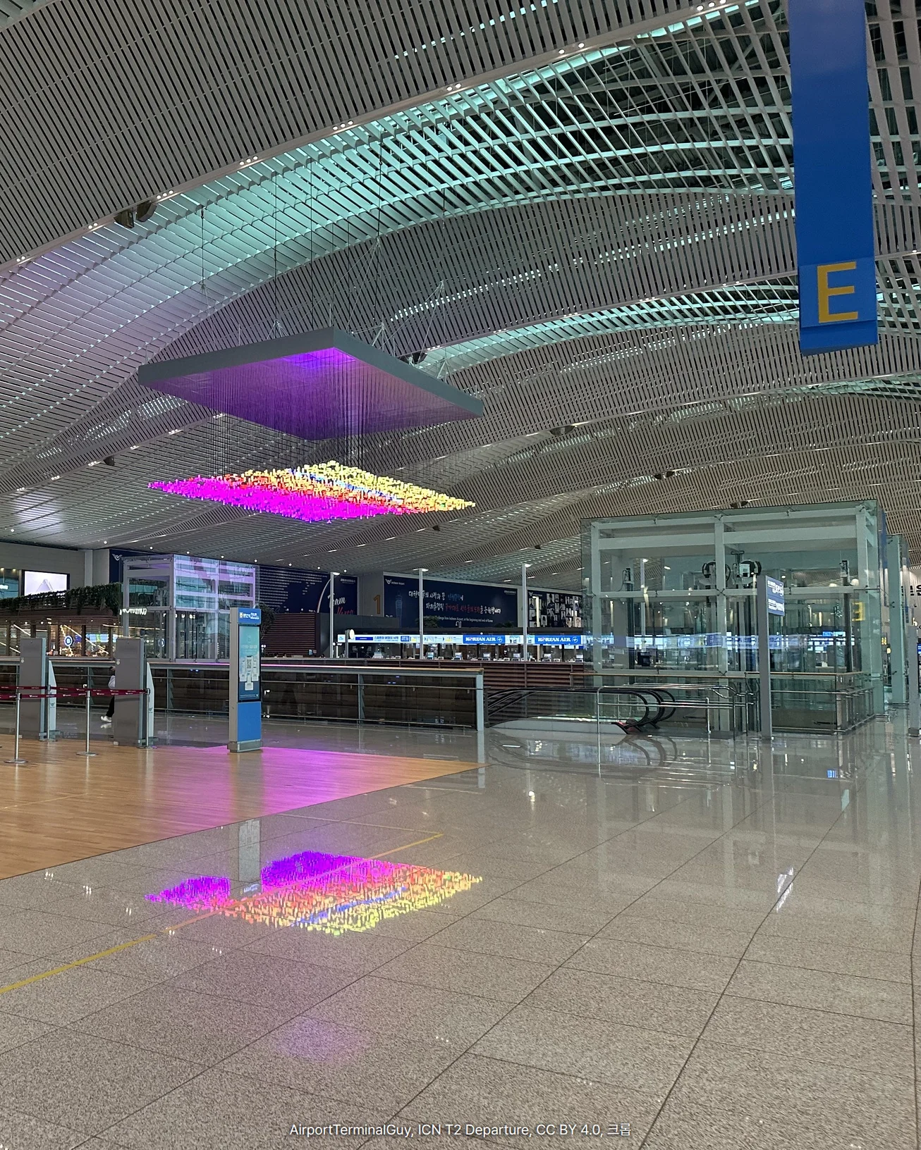인천공항 제2여객터미널 출국장의 높은 곡면 천장에 E 구역 표지와 빛 조형물이 매달려 있고, 그 아래 체크인 카운터와 조형물의 빛이 비친 바닥이 보이는 사진입니다