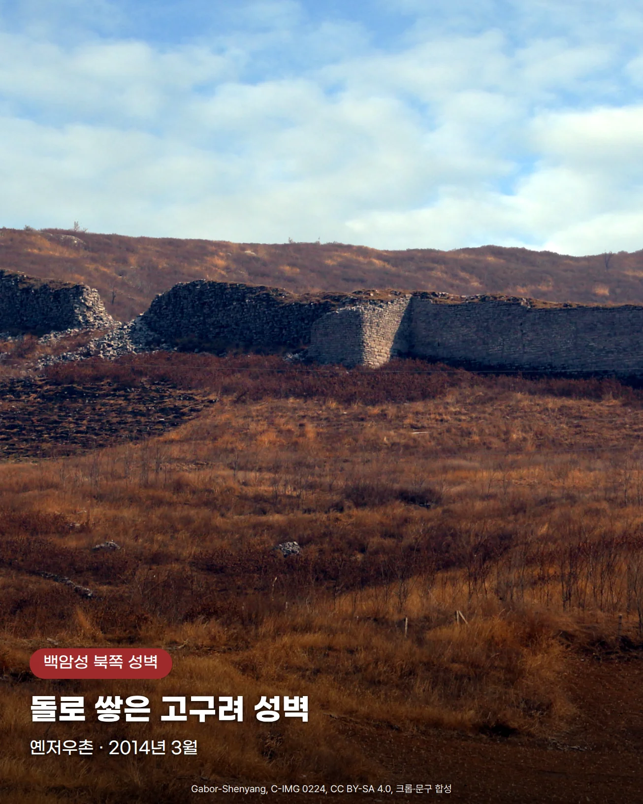 마른 갈색 풀이 덮인 언덕 위로 돌로 쌓은 성벽이 가로로 이어지고, 성벽 중간중간 바깥으로 튀어나온 모서리가 있는 사진. 위쪽은 구름 낀 파란 하늘이다. 아래쪽에 '백암성 북쪽 성벽' 배지와 '돌로 쌓은 고구려 성벽', '옌저우촌 · 2014년 3월' 문구가 있다.