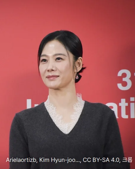 제31회 부산국제영화제 실낙원 기자회견 포토월에 선 김현주. 붉은 배경 앞에서 레이스 깃이 달린 짙은 회색 니트를 입고 옅게 미소 지으며 비스듬히 앞을 보고 있습니다