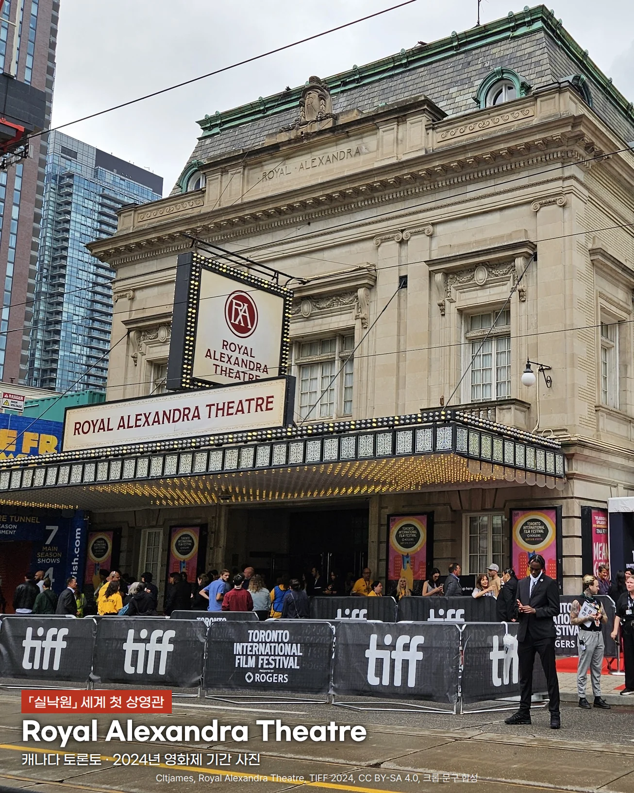 토론토국제영화제 기간의 Royal Alexandra Theatre 외관 사진. 흐린 하늘 아래 베이지색 석조 극장 건물 앞에 ROYAL ALEXANDRA THEATRE 간판과 RA 로고 간판이 달려 있고, 길가에는 tiff 로고가 찍힌 펜스와 레드카펫 주변에 사람들이 모여 있으며, 아래쪽 길 위에 실낙원 세계 첫 상영관, Royal Alexandra Theatre, 캐나다 토론토 2024년 영화제 기간 사진이라는 문구가 있습니다