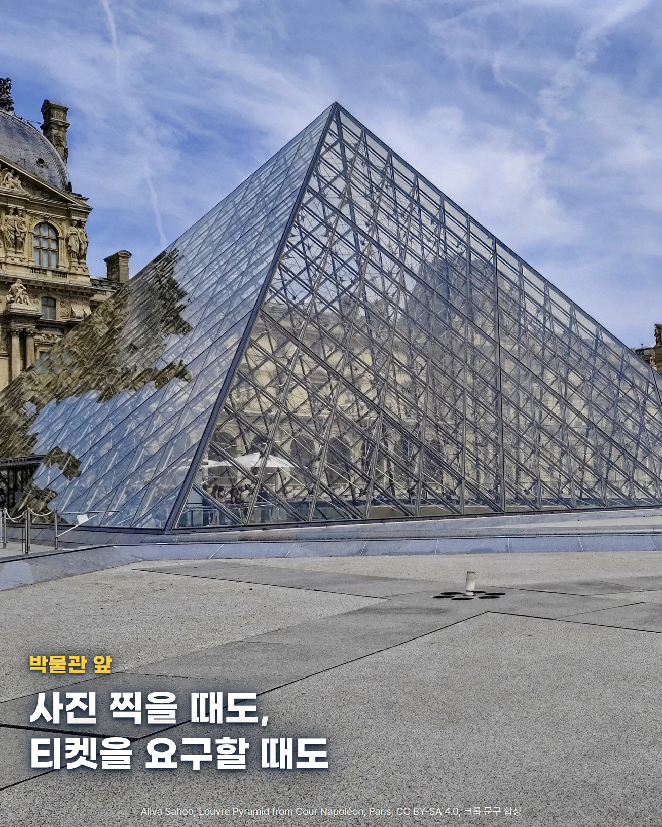 푸른 하늘 아래 유리 피라미드가 화면 위쪽 절반을 채우고, 왼쪽에 루브르 궁 건물 일부, 아래쪽에 넓은 회색 광장 바닥이 보이는 사진입니다. 아래쪽 광장 바닥 위에 박물관 앞, 사진 찍을 때도 티켓을 요구할 때도라는 문구가 있습니다.