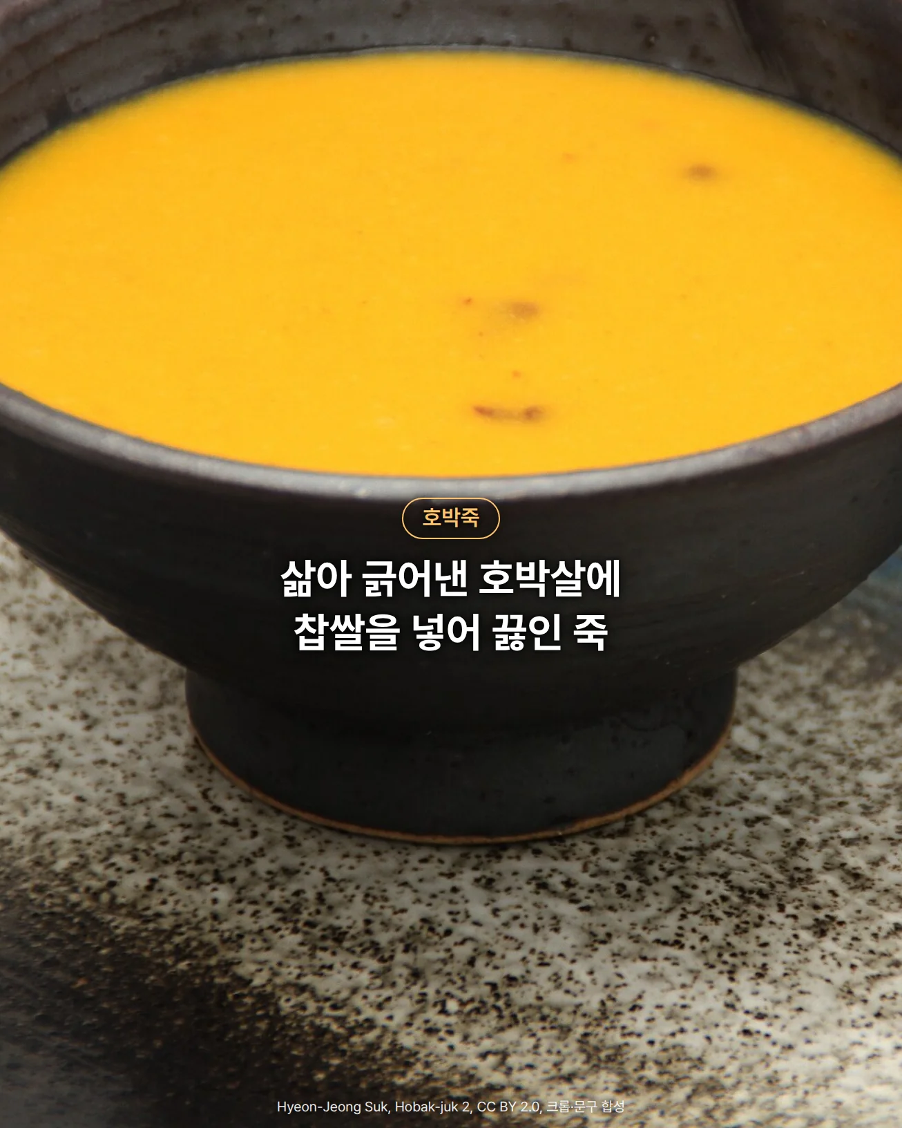 검은 도자기 그릇에 담긴 매끈하고 진한 주황색 호박죽을 가까이에서 찍은 사진으로, 그릇이 푸른 결과 흰 점무늬가 있는 접시 위에 놓여 있고, 그릇 몸통 위에 '호박죽' 배지와 '삶아 긁어낸 호박살에 찹쌀을 넣어 끓인 죽' 문구가 있습니다
