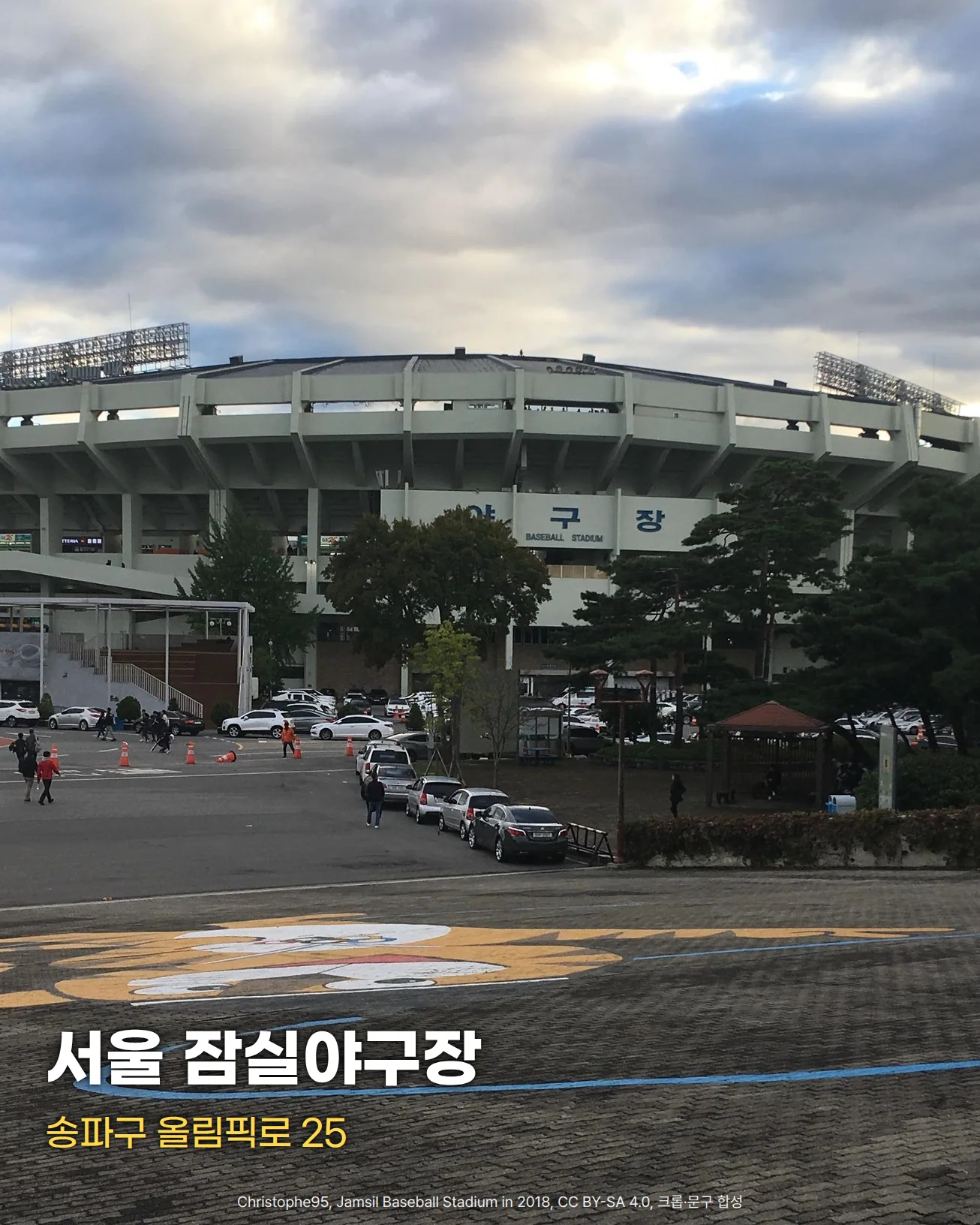 흐린 하늘 아래 넓은 광장 건너편에 둥근 콘크리트 외벽의 잠실야구장이 보이는 사진입니다. 정면에 '야구장 BASEBALL STADIUM' 글자가 보이고, 구장 앞에는 주차된 차량과 걸어가는 사람들, 오른쪽에는 소나무가 있습니다. 아래쪽에 '서울 잠실야구장'과 '송파구 올림픽로 25' 문구가 있습니다.