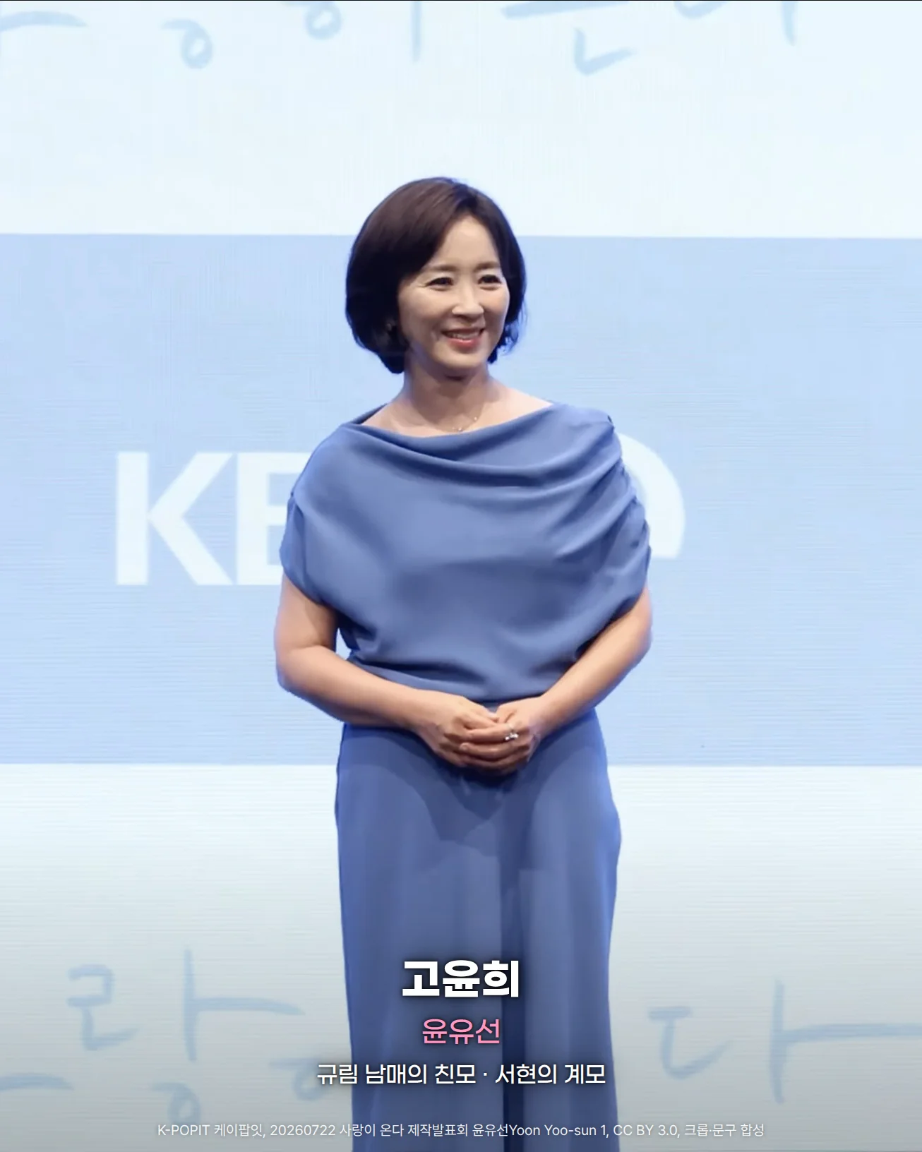 사랑이 온다 제작발표회 무대에서 'KBS'와 '사랑이 온다' 글자가 있는 하늘색 배경 앞에 파란 블라우스와 바지를 입은 윤유선이 두 손을 모으고 웃으며 선 사진. 아래쪽에 '고윤희', '윤유선', '규림 남매의 친모 · 서현의 계모' 문구가 있습니다.