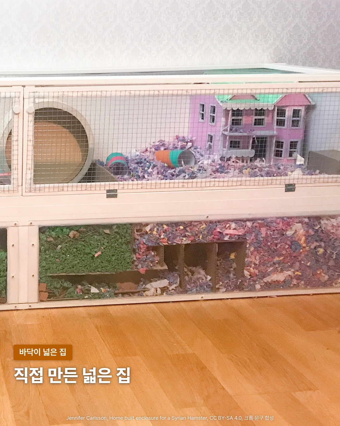 마루 위에 놓인 흰 나무 틀의 가로로 긴 2단 햄스터 집을 정면에서 찍은 사진입니다. 위칸 철망 너머로 나무 쳇바퀴와 분홍색 집 모양 은신처, 색색의 종이 베딩이 보이고, 아래칸 유리 너머로는 초록 베딩과 색색의 종이 베딩이 깊게 쌓인 사이에 칸막이로 나뉜 은신처가 보입니다. 아래쪽 마루 위에 '직접 만든 넓은 집'이라는 글자가 있습니다.