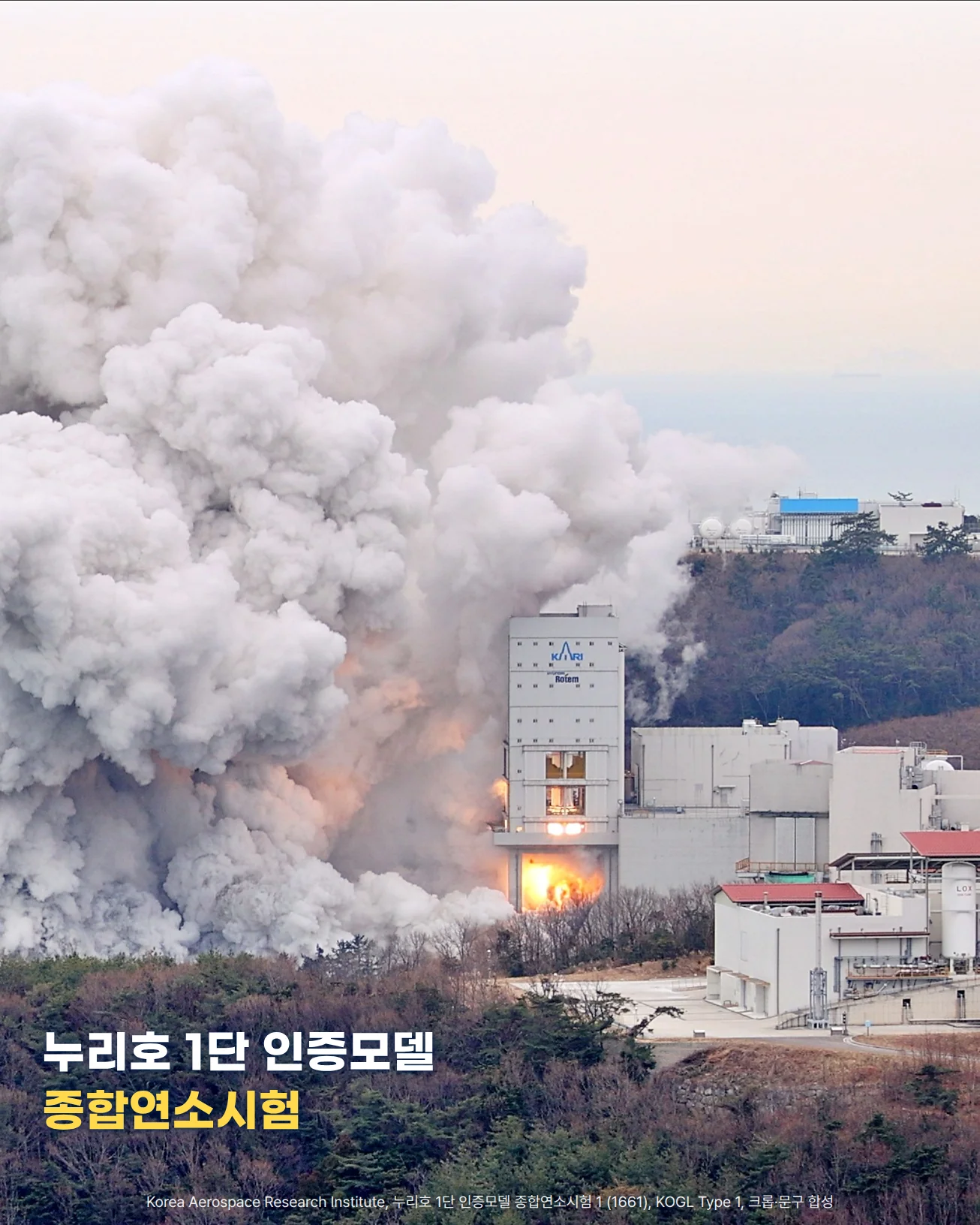 바닷가 언덕 위 시험대에서 누리호 1단 인증모델 종합연소시험이 진행되는 모습. 시험대 아래쪽에서 주황색 화염이 보이고 거대한 흰 연기 구름이 바다 쪽 하늘로 피어오릅니다. 아래쪽 숲 위에 '누리호 1단 인증모델 종합연소시험' 문구가 놓여 있습니다