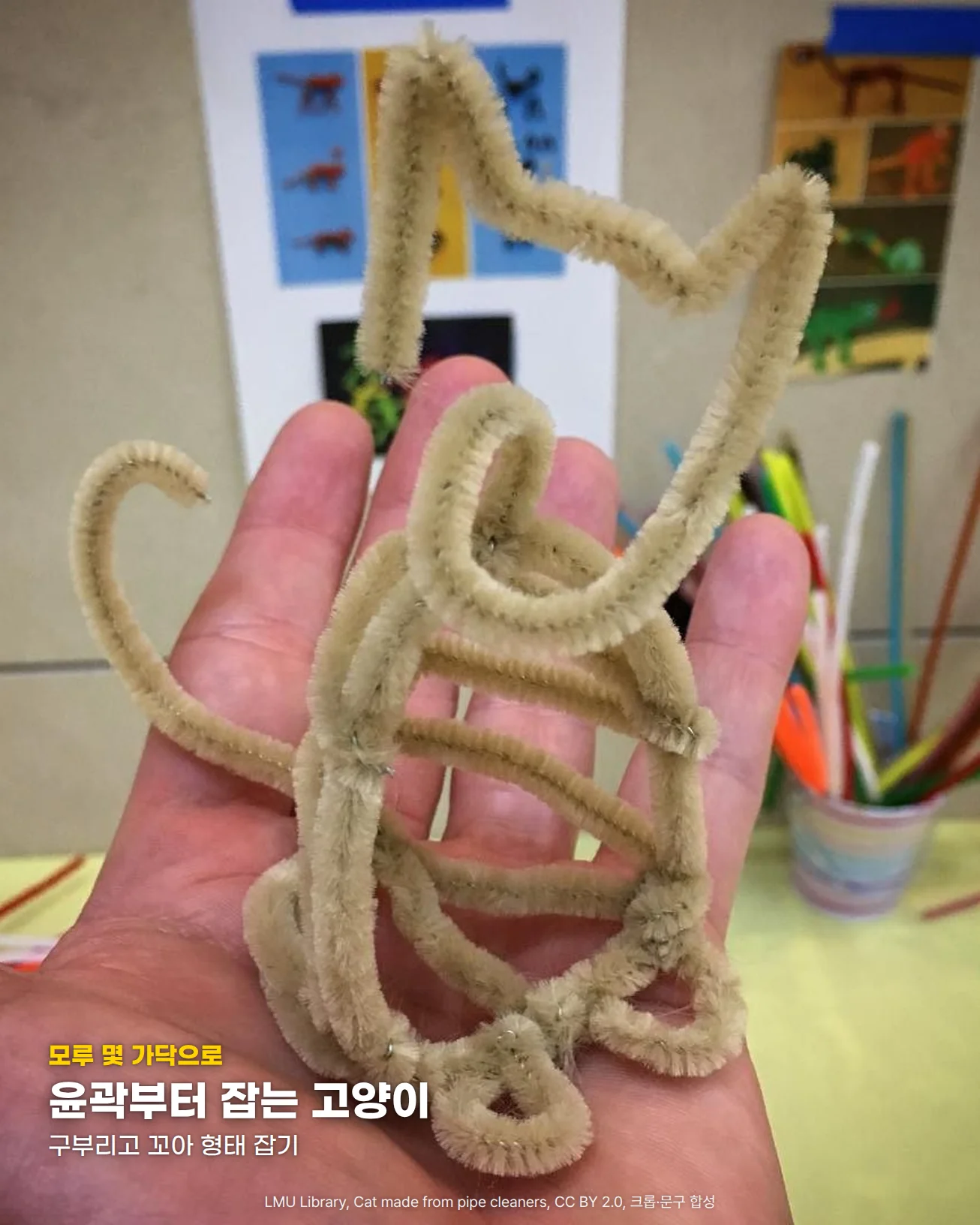 손바닥 위에 베이지색 모루 몇 가닥을 구부리고 꼬아 귀, 얼굴, 몸통, 꼬리의 윤곽만 선으로 만든 고양이가 서 있는 사진입니다. 뒤쪽 탁자 위 컵에는 빨강, 주황, 초록, 파랑 모루가 꽂혀 있습니다. 아래쪽에 '모루 몇 가닥으로' 문구와 '윤곽부터 잡는 고양이' 제목, '구부리고 꼬아 형태 잡기' 문구가 있습니다.