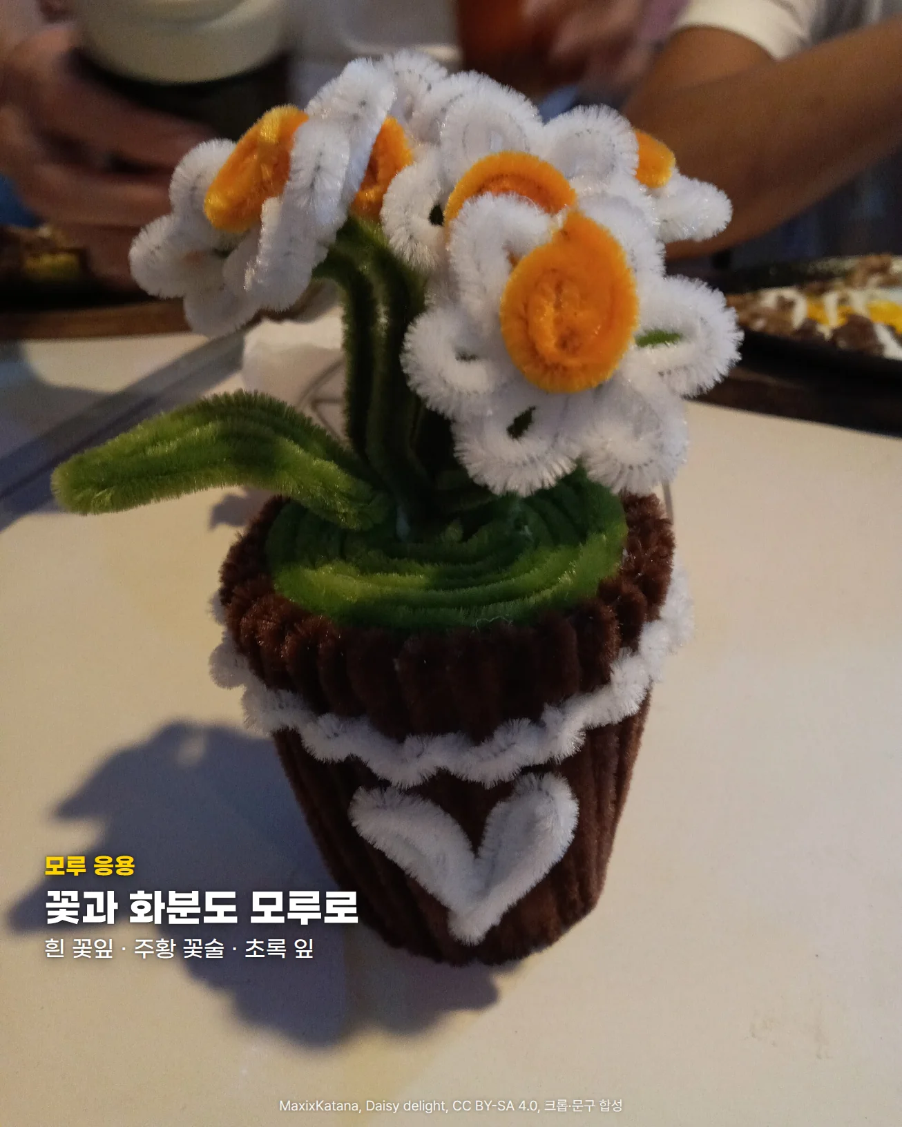 흰 탁자 위에 갈색 모루를 감아 만든 화분이 있고, 흰 모루 꽃잎과 주황 모루 꽃술로 만든 데이지 여러 송이와 초록 모루 잎이 꽂혀 있는 사진입니다. 화분 둘레에는 흰 모루 물결 장식과 하트가 붙어 있습니다. 왼쪽 아래에 '모루 응용' 문구와 '꽃과 화분도 모루로' 제목, '흰 꽃잎 · 주황 꽃술 · 초록 잎' 문구가 있습니다.