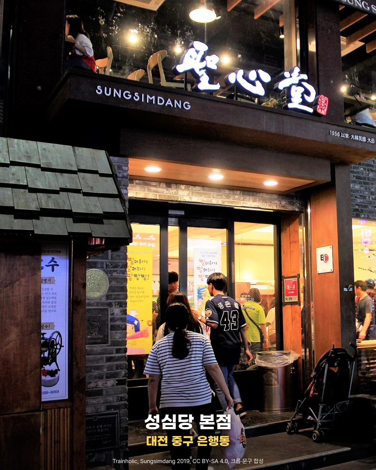밤에 조명이 켜진 대전 은행동 성심당 본점 입구. 위쪽에 '聖心堂' 간판과 'SUNGSIMDANG' 글자가 보이고, 사람들이 유리문 안으로 들어간다. 사진 아래쪽에 '성심당 본점'과 '대전 중구 은행동' 문구