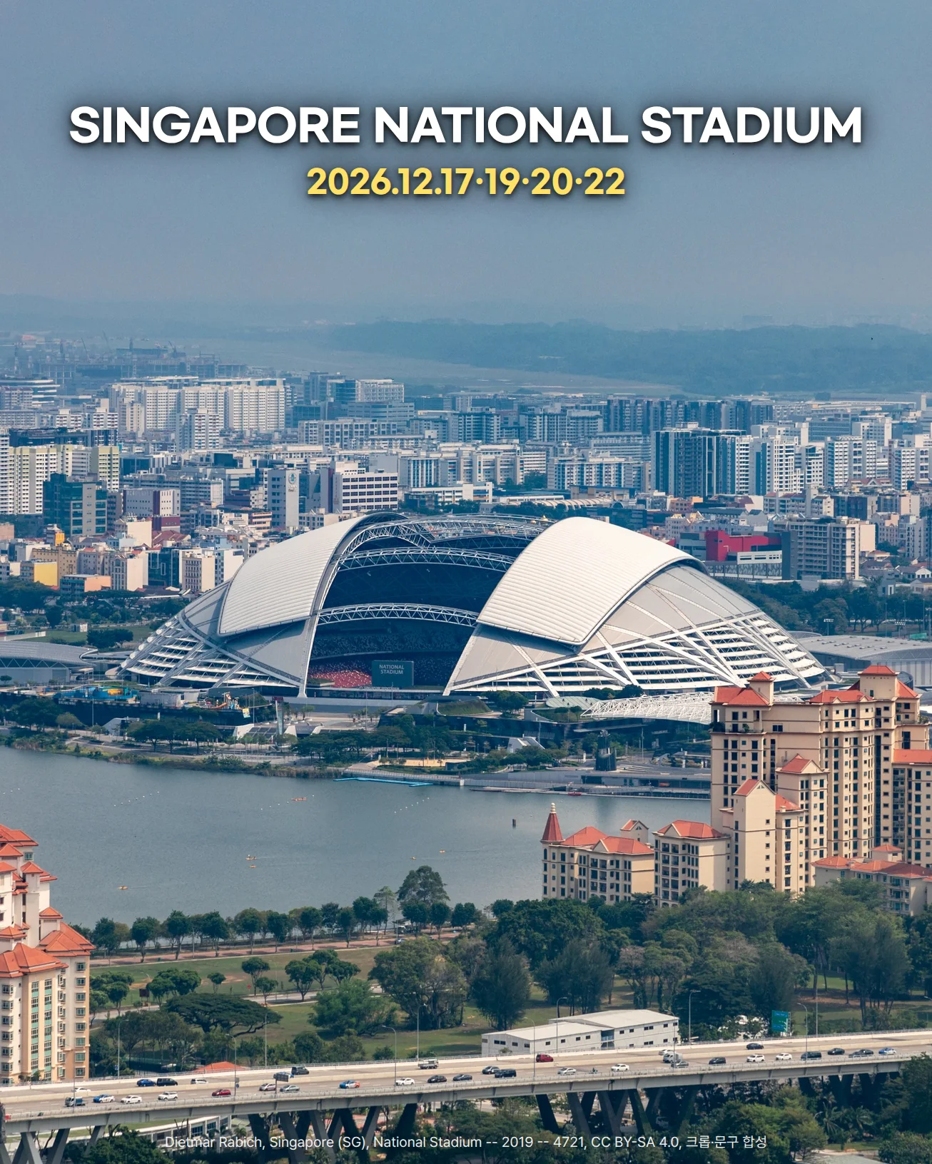 높은 곳에서 내려다본 싱가포르 National Stadium 사진입니다. 칼랑 강 너머에 흰 돔 지붕 경기장이 있고, 그 뒤로 도심의 건물들이 이어지며, 앞쪽에는 강가의 나무와 고층 아파트, 고가도로가 있습니다. 위쪽 하늘에 'SINGAPORE NATIONAL STADIUM'과 '2026.12.17·19·20·22' 문구가 있습니다.
