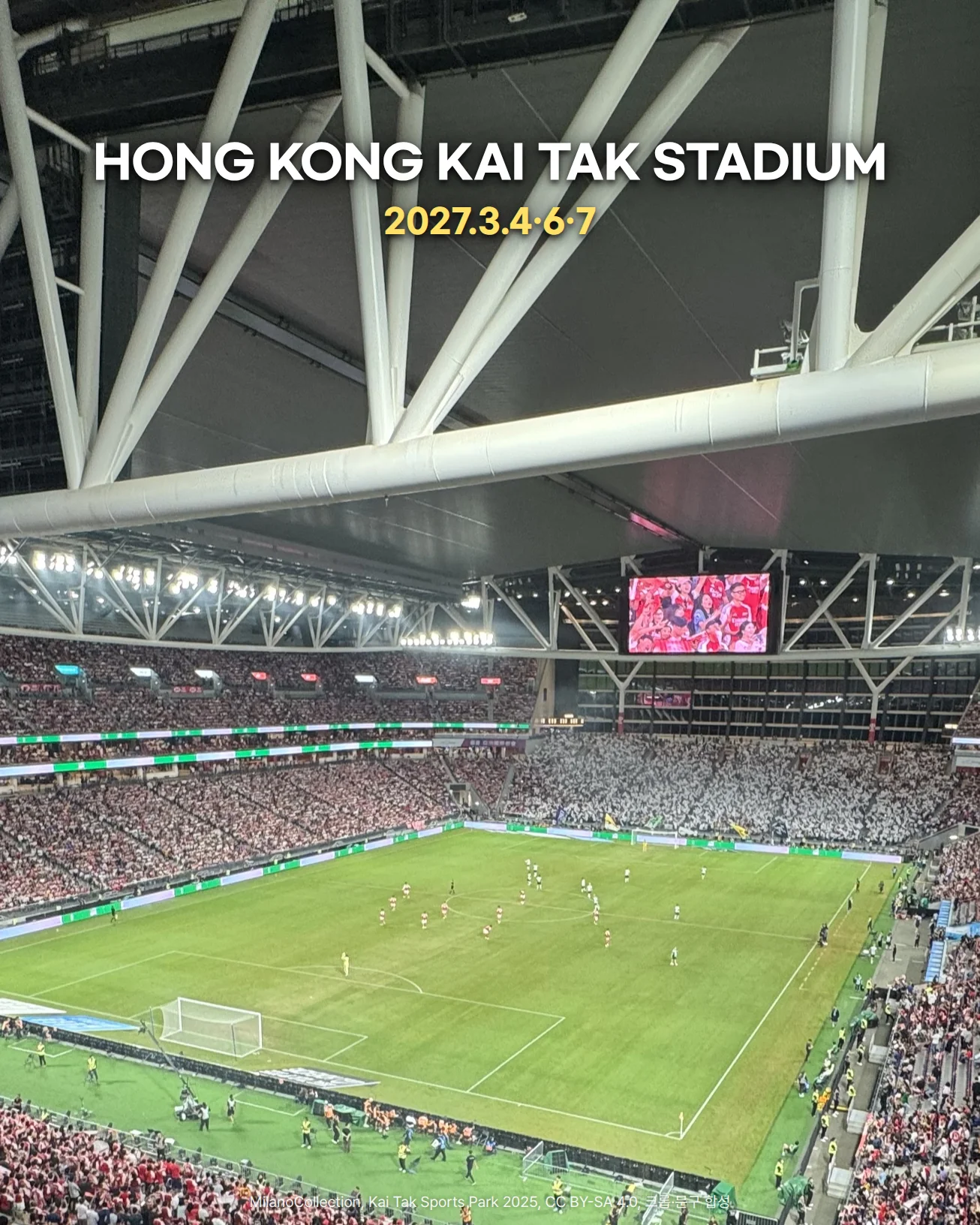 홍콩 Kai Tak Stadium 안을 위층 관중석에서 내려다본 사진입니다. 흰 철골 구조의 지붕 아래 대형 전광판이 달려 있고, 조명이 켜진 잔디 그라운드를 관중이 가득 찬 관중석이 둘러싸고 있습니다. 위쪽 지붕 아래에 'HONG KONG KAI TAK STADIUM'과 '2027.3.4·6·7' 문구가 있습니다.