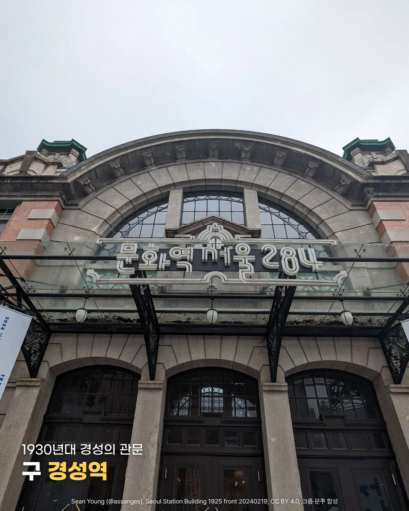 흐린 하늘 아래 아치형 큰 창과 붉은 벽돌 벽, 초록 지붕 장식이 있는 구 경성역 건물 정면을 올려다본 사진입니다. 유리 차양 위에 '문화역서울284' 간판이 있고 아래에 아치형 출입문 세 개가 보이며, 아래쪽 어두운 출입문 위에 '1930년대 경성의 관문'과 '구 경성역'이라는 글자가 놓여 있습니다