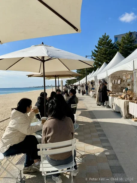 강릉커피거리 바닷가 쪽 보행로에 놓인 파라솔과 야외 테이블에서 바다를 바라보며 커피를 마시는 방문객, 그 옆으로 공예품과 생활소품을 진열한 흰 천막 가판이 이어지는 낮 장면