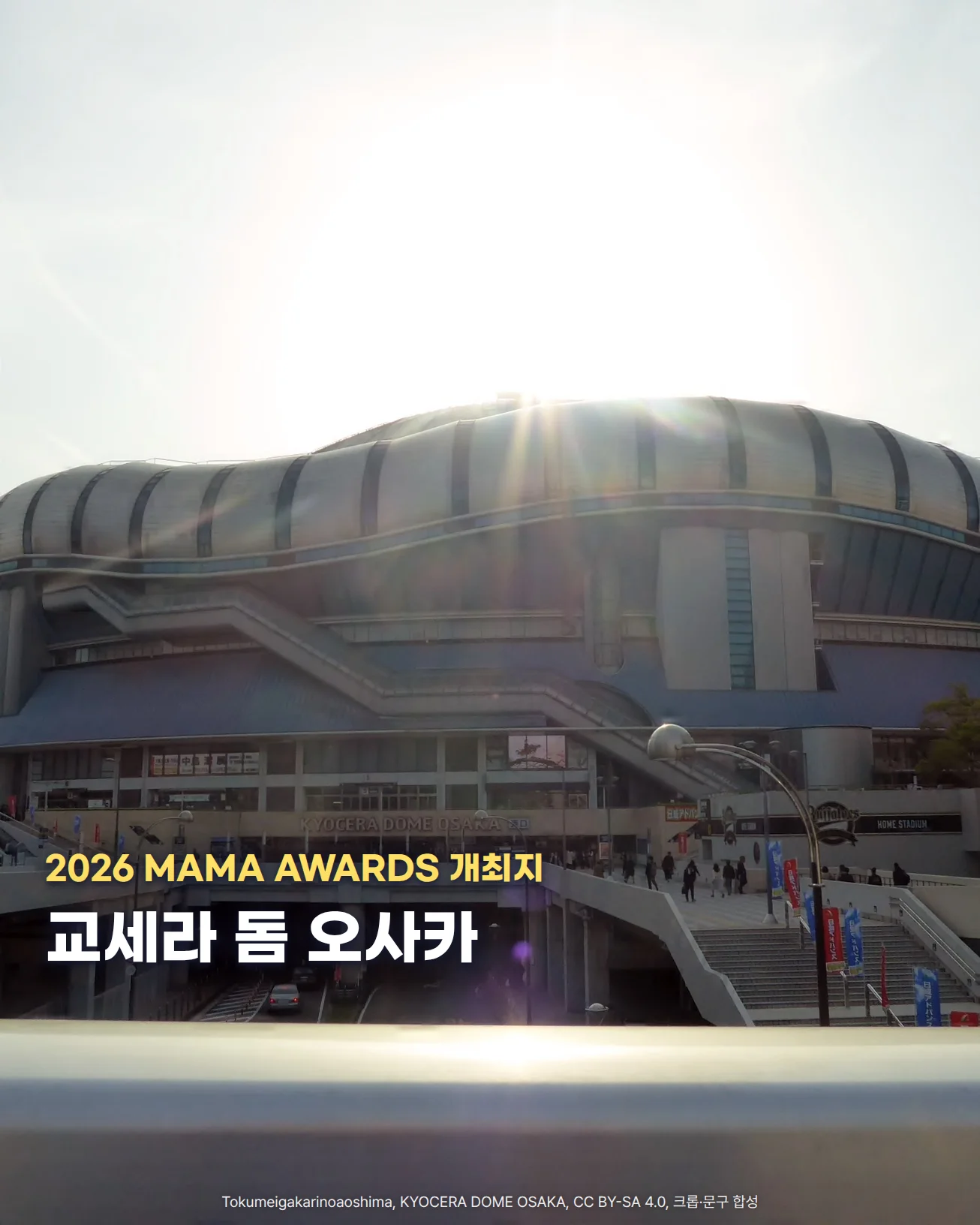 역광 속 하늘 아래 물결 모양 지붕의 교세라 돔 오사카 외관과 그 앞의 보행 데크, 계단을 오가는 사람들이 보이는 사진입니다. 왼쪽 아래에 교세라 돔 오사카, 2026 MAMA AWARDS 개최지라는 문구가 있습니다.