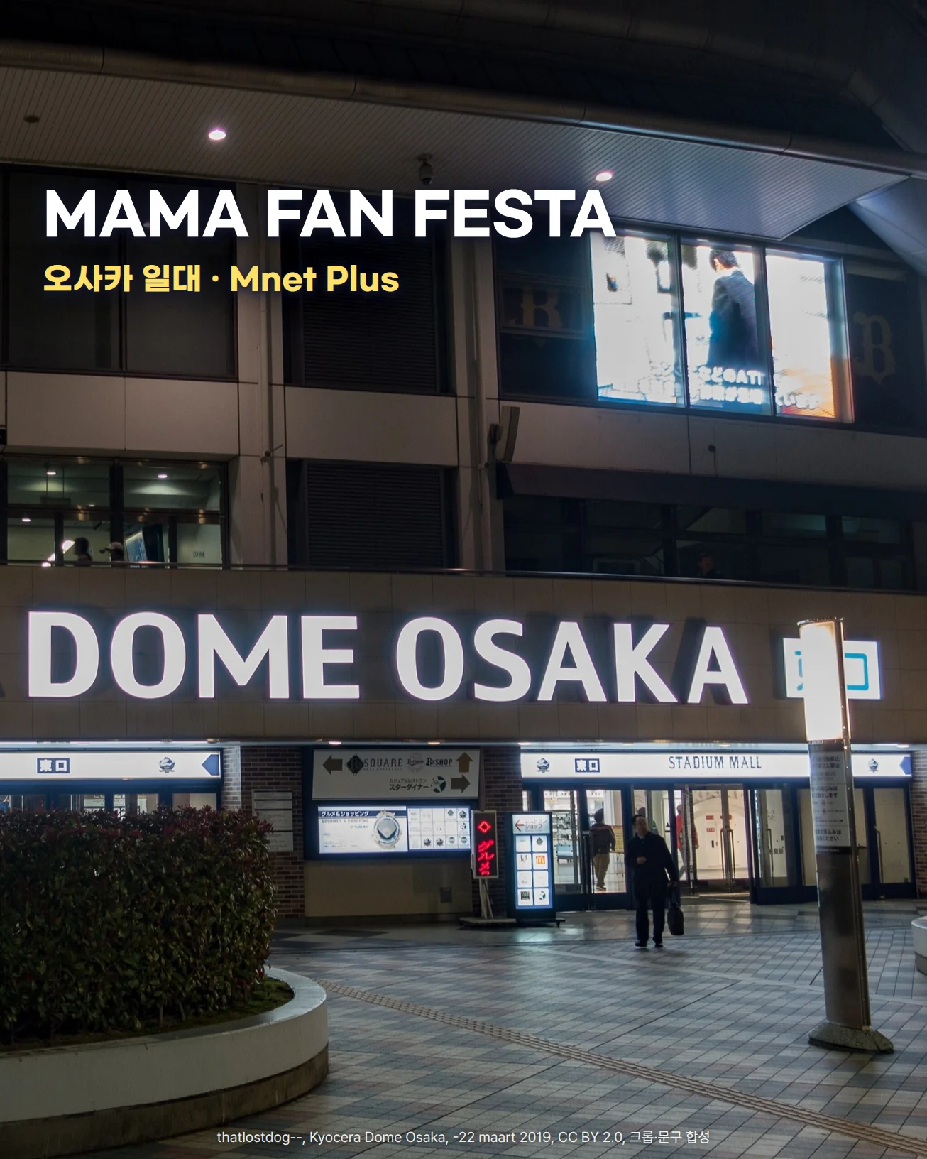 밤에 불이 켜진 DOME OSAKA 간판 아래로 STADIUM MALL 東口 입구와 화단, 타일 보도가 보이고 입구 앞을 한 사람이 지나가는 사진입니다. 왼쪽 위 어두운 유리창 위에 MAMA FAN FESTA, 오사카 일대 · Mnet Plus라는 문구가 있습니다.