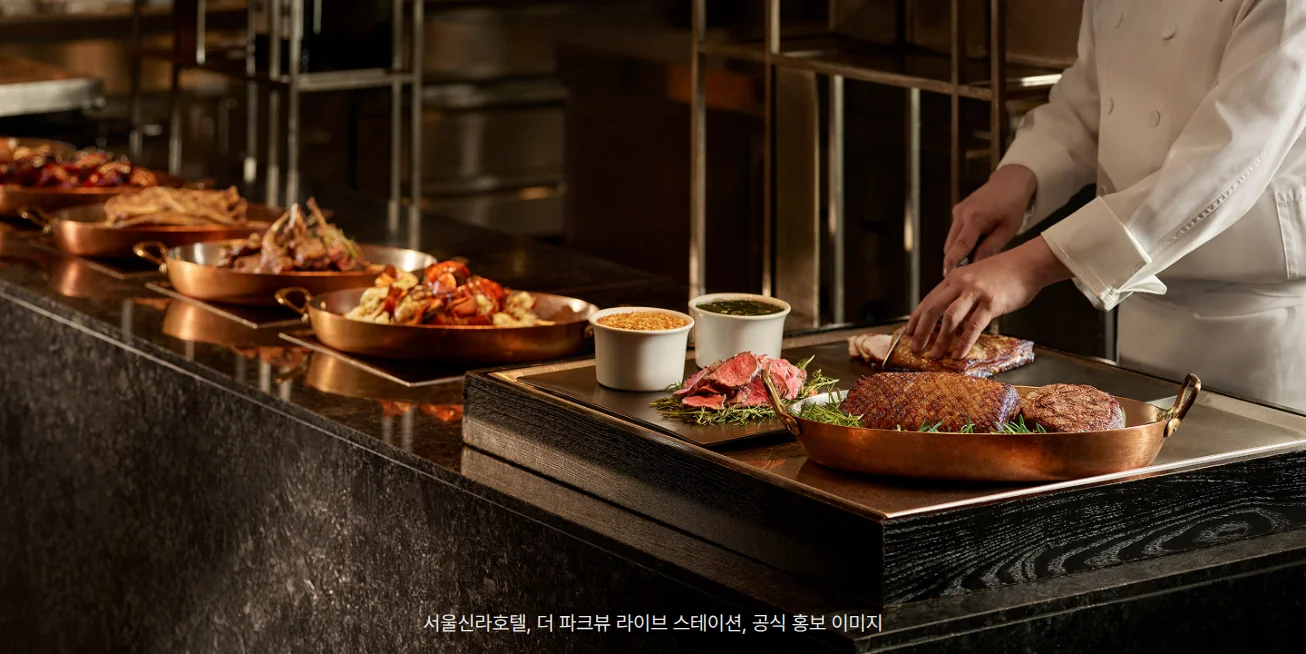 검은 대리석 조리대 위에 구리 팬이 줄지어 놓이고, 흰 조리복을 입은 셰프가 오른쪽에서 칼로 구운 고기를 자르는 더 파크뷰 라이브 스테이션 사진. 앞쪽 구리 팬에는 스테이크와 로즈메리, 그 옆 접시에는 얇게 썬 고기와 소스 두 그릇이 있고, 뒤쪽 팬에는 랍스터와 다른 요리가 담겨 있습니다