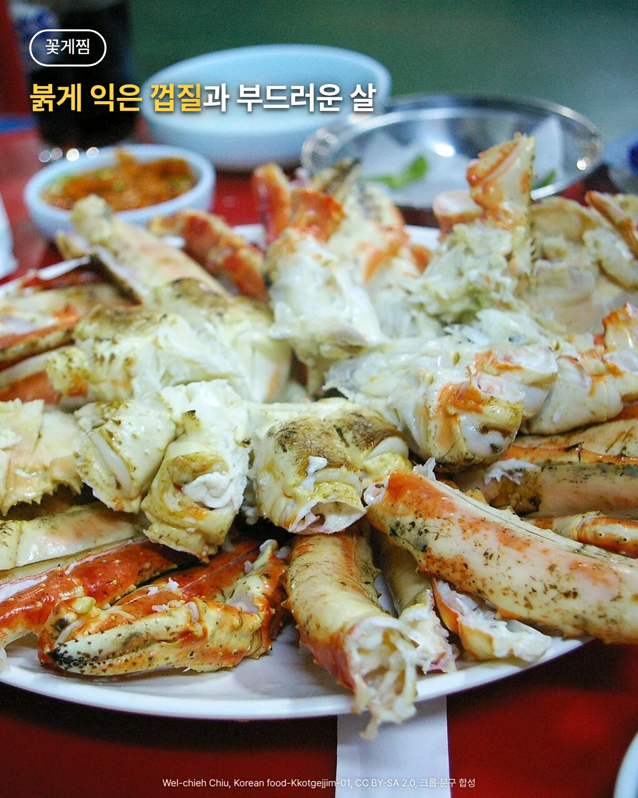 붉은 식탁 위 흰 접시에 쪄 낸 꽃게찜이 수북이 담긴 사진. 등딱지를 분리하고 반으로 자른 몸통과 다리 토막마다 붉게 익은 껍질과 흰 살이 드러나 있고, 뒤로 양념장 종지와 빈 그릇이 보인다. 위쪽에 '꽃게찜', '붉게 익은 껍질과 부드러운 살' 문구가 들어가 있다