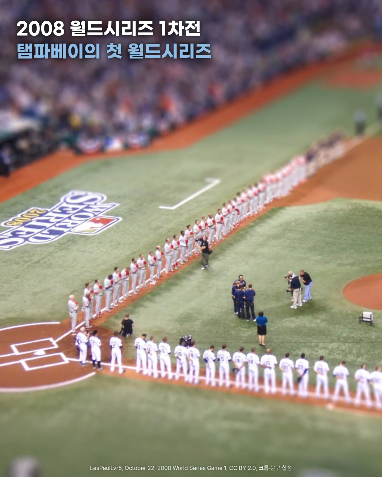 2008년 10월 22일 트로피카나 필드 월드시리즈 1차전 개막 행사를 위쪽 관중석에서 내려다본 사진으로, 양 팀 선수단이 홈플레이트에서 뻗은 두 파울라인을 따라 한 줄로 서 있고 잔디에 2008 WORLD SERIES 로고가 그려져 있으며, '2008 월드시리즈 1차전', '탬파베이의 첫 월드시리즈' 문구가 있는 이미지