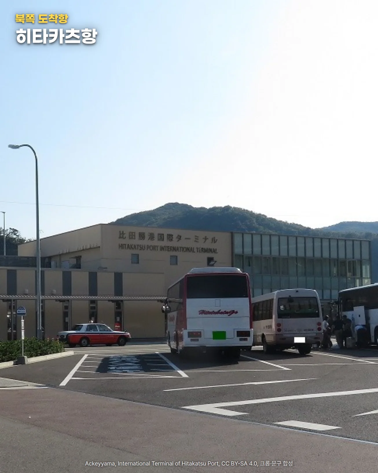 맑은 날 히타카츠항 국제터미널 앞. 낮은 베이지색 건물 위에 '比田勝港国際ターミナル'와 'HITAKATSU PORT INTERNATIONAL TERMINAL' 간판이 있고, 앞 광장에 관광버스와 택시가 서 있으며 뒤로 초록 산이 보입니다. 왼쪽 위에 '북쪽 도착항' 라벨과 '히타카츠항' 글자가 있습니다