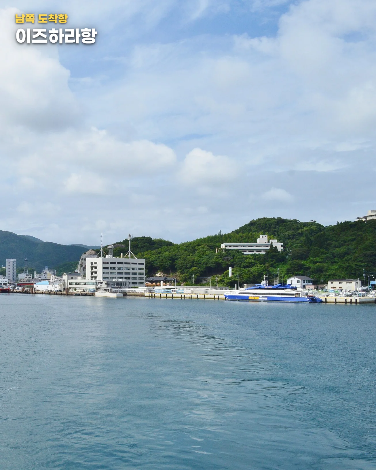 흐린 하늘 아래 바다에서 바라본 이즈하라항. 초록 산으로 둘러싸인 항구 안쪽에 시가지와 흰 건물들이 있고, 오른쪽 부두에 파란색 고속선이 정박해 있습니다. 왼쪽 위에 '남쪽 도착항' 라벨과 '이즈하라항' 글자가 있습니다