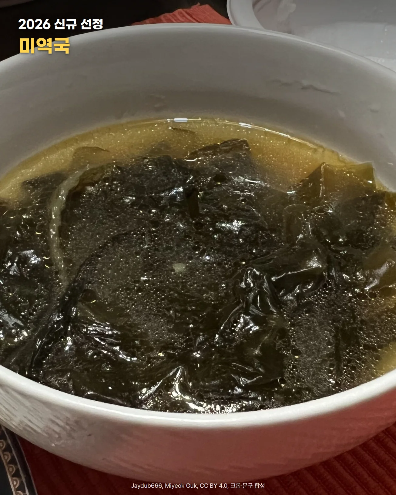 흰 그릇 가득 미역이 담긴 미역국을 가까이에서 본 사진. 붉은 식탁 매트 위에 그릇이 놓였다. 사진 위에 '2026 신규 선정, 미역국' 문구