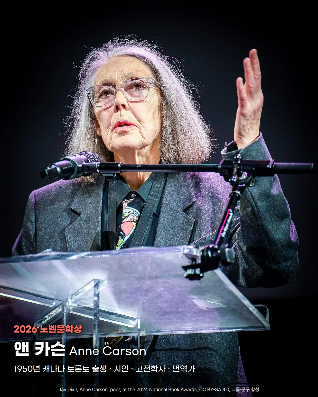 어두운 무대에서 투명 강연대와 마이크 앞에 서서 오른손을 들어 올리며 말하는 앤 카슨의 상반신 사진입니다. 은발에 안경을 끼고 회색 재킷을 입었습니다. 왼쪽 아래에 '2026 노벨문학상', '앤 카슨 Anne Carson', '1950년 캐나다 토론토 출생 · 시인 · 고전학자 · 번역가' 글자가 있습니다