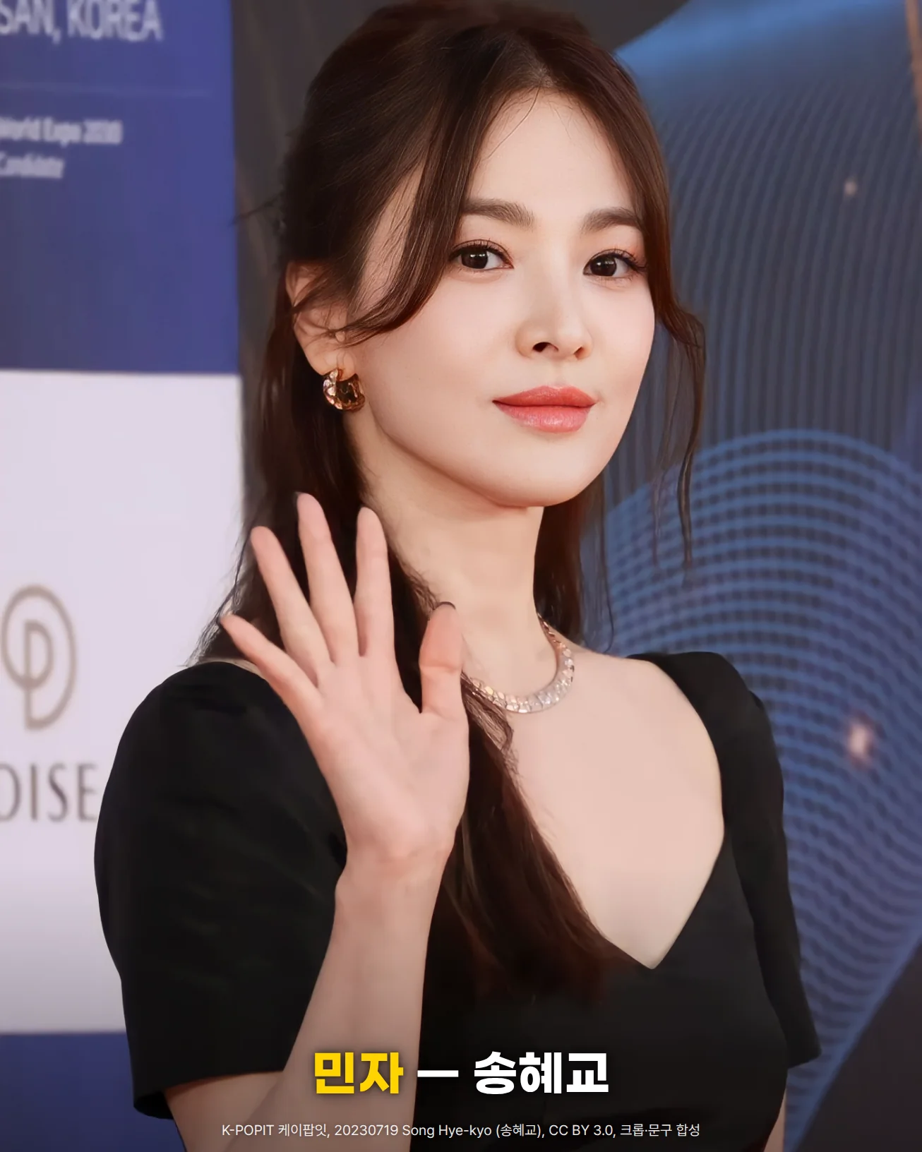 제2회 청룡시리즈어워즈 레드카펫에서 긴 머리를 뒤로 넘기고 검은 드레스와 목걸이 차림으로 오른손을 들어 인사하는 송혜교의 상반신 사진. 아래쪽에 '민자 — 송혜교' 문구가 있습니다
