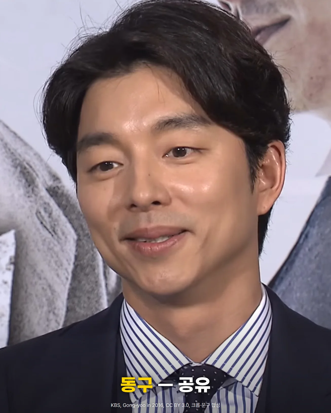 짙은 남색 정장에 줄무늬 셔츠와 넥타이 차림으로 미소 짓는 공유의 얼굴과 상반신 사진. 뒤로 흰 배경의 포스터가 보이고, 아래쪽에 '동구 — 공유' 문구가 있습니다
