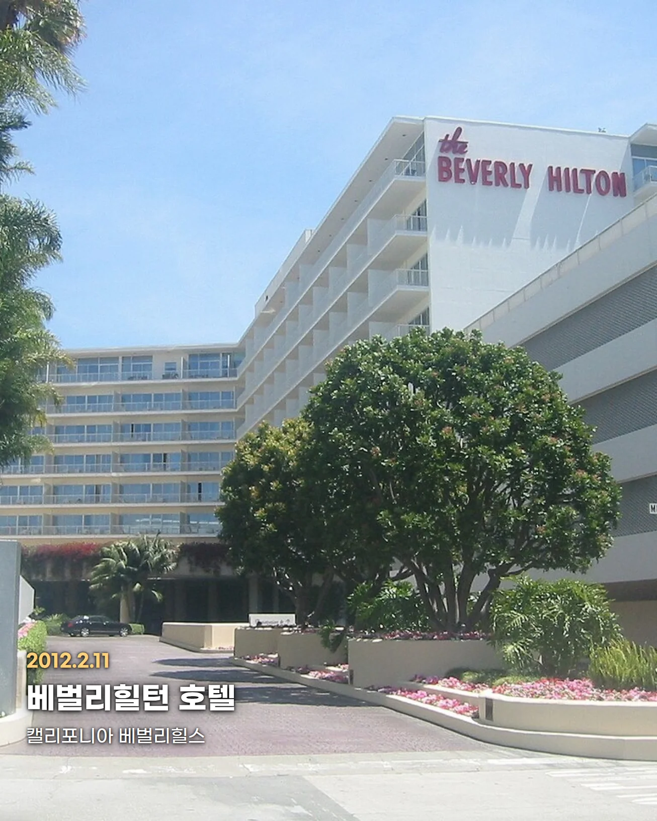캘리포니아 베벌리힐스의 베벌리힐턴 호텔 외관. 맑은 하늘 아래 흰색 객실 타워가 서 있고, 오른쪽 건물 위에 붉은 글씨의 'the BEVERLY HILTON' 간판이 있으며, 앞쪽 진입로를 따라 야자수와 둥근 나무, 꽃밭이 이어집니다. 왼쪽 아래에 '2012.2.11', '베벌리힐턴 호텔', '캘리포니아 베벌리힐스' 글자가 있습니다