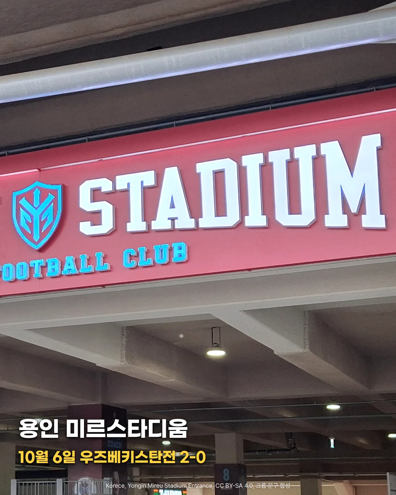 용인 미르스타디움 입구 위 붉은 간판에 흰 글자 STADIUM과 하늘색 엠블럼이 있고 그 아래로 천장과 입장 게이트 위쪽이 보이는 사진에 '용인 미르스타디움', '10월 6일 우즈베키스탄전 2-0' 문구가 있는 이미지
