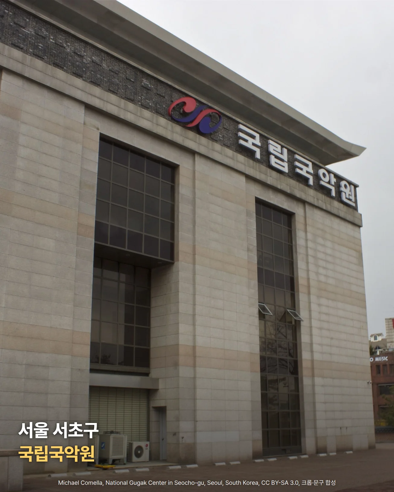 흐린 하늘 아래 돌판으로 마감한 높은 건물의 정면을 비스듬히 올려다본 사진입니다. 건물 위쪽 처마 아래에 붉은색과 파란색의 태극 모양 상징과 '국립국악원' 글자 간판이 붙어 있고, 벽면 가운데에는 세로로 긴 유리창이 나 있습니다. 왼쪽 아래에 '서울 서초구 국립국악원'이라는 글자가 놓여 있습니다.