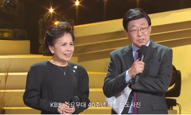 가요무대 40주년 특집 무대에서 검은 옷을 입은 이미자가 마이크를 들고 미소 짓고, 그 옆에서 정장 차림의 김동건 아나운서가 마이크를 들고 말하는 장면입니다. 뒤로 노란 조명이 비친 계단식 무대가 보입니다.
