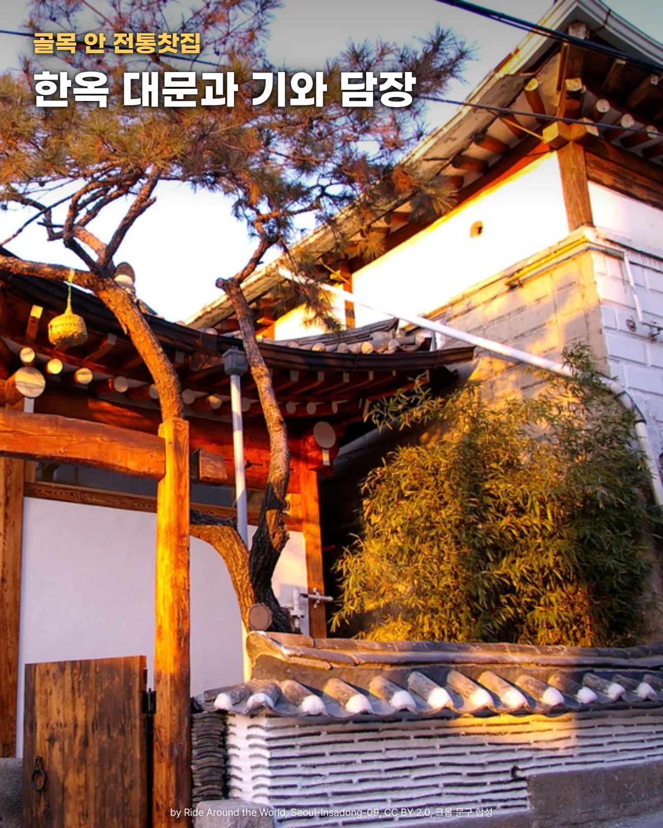 기와를 얹은 한옥 처마 아래 나무 대문과 굵은 나무 기둥이 서 있고, 그 앞으로 소나무 줄기가 비스듬히 뻗어 있습니다. 오른쪽에는 기와를 올린 흰 담장과 대나무, 흰 벽의 건물이 저녁 햇빛을 받고 있습니다. 위쪽에 골목 안 전통찻집, 한옥 대문과 기와 담장이라는 문구가 있습니다